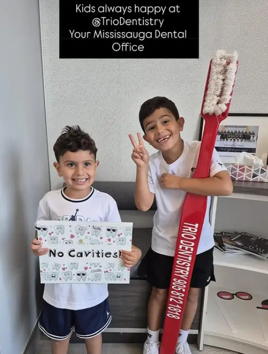 Two smiling boys stand in a dental office; one holds a “No Cavities!” sign, and the other holds a giant toothbrush with “TRIODENTISTRY 905-812-1818” printed on it.