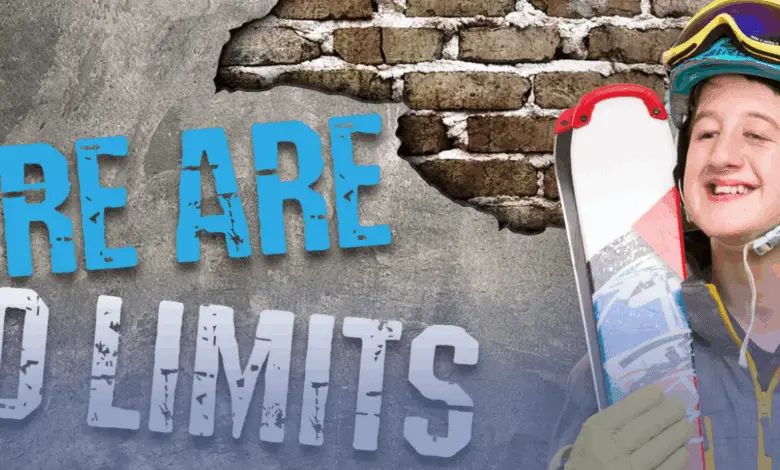 Smiling skier in a helmet and goggles holds skis beside large text on a brick wall that reads: “There Are No Limits.”