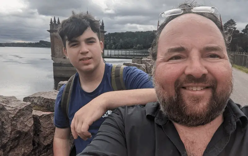 My son and I after a long hike. outdoors on a cloudy day. standing on a stone walkway beside a large body of water with a historic-looking tower structure and railings in the background. One person is smiling at the camera, while the other stands close by with a neutral expression and a backpack. The mood feels relaxed and casual, with natural scenery behind them.