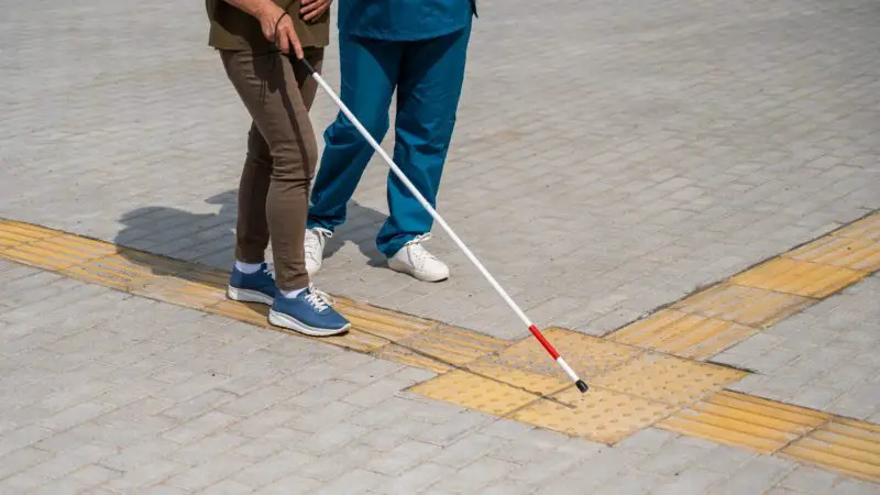 Two people walking on a sidewalk with yellow Tenji blocks; one uses a white cane to follow the tactile path.