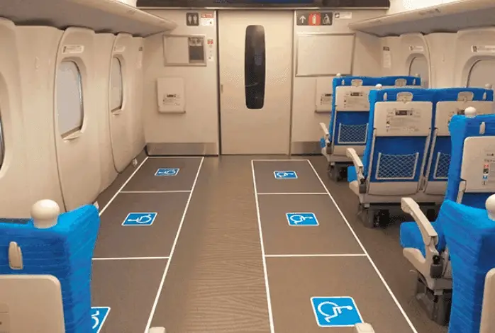Wheelchair spaces with accessibility symbols marked on the floor inside a Shinkansen train car. Blue passenger seats line the right side.
