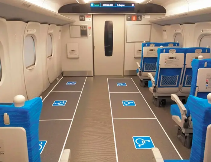 Wheelchair spaces with accessibility symbols marked on the floor inside a Shinkansen train car. Blue passenger seats line the right side.