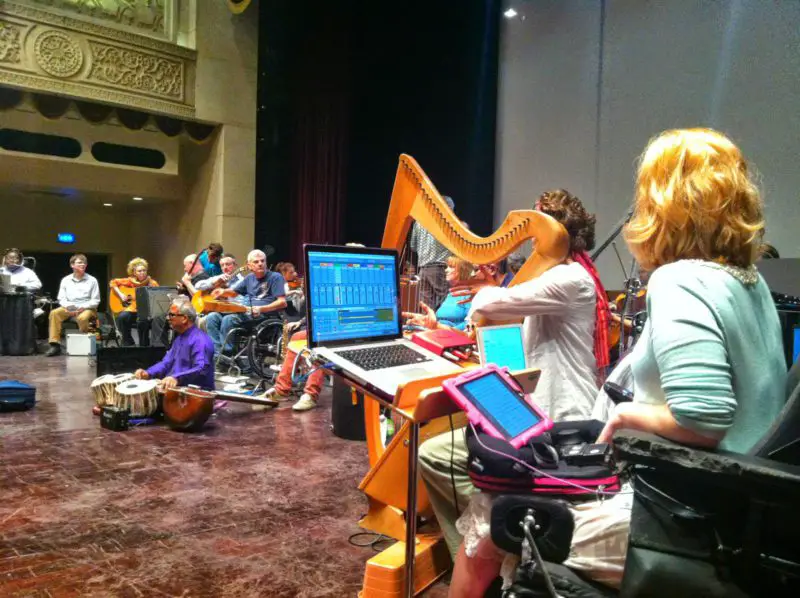 Inclusive ensemble rehearsal on a theatre stage, with wheelchair users and non-disabled musicians using acoustic and digital instruments.