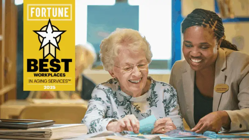 A staff member sits beside an older woman at a table, both smiling as they look at papers together. A yellow graphic reads “Fortune Best Workplaces in Aging Services 2025.”