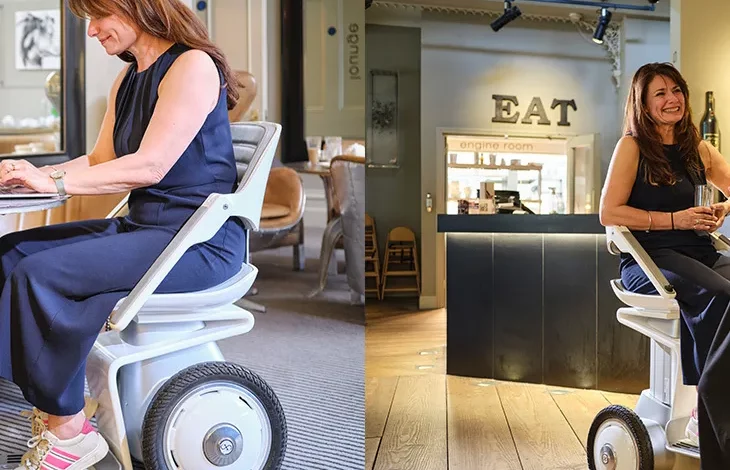 Two photos show a woman seated on a white self-balancing, two-wheel mobility device indoors. In the first image, she sits at a table working on a laptop. In the second, she is raised to standing height at a bar counter, smiling and holding a drink. The device has large central wheels and a compact base, allowing her to interact at eye level in a café setting.
