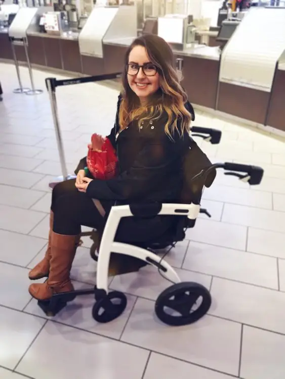 Smiling woman sitting on a rollator with a seat inside a cinema lobby, holding a snack bag while waiting near the counter.