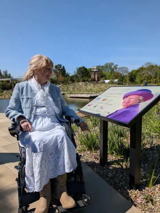 Clare using a wheelchair reads an information board about Queen Elizabeth II garden beside a pond in the garden. 