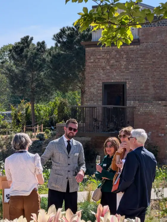 Group of people chat beside planting near the garden’s water tower, holding drinks during an opening event.