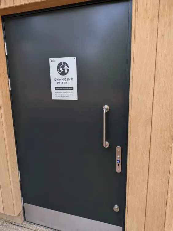Door to a Changing Places toilet with RADAR key access sign and grab handle on a wooden-clad building.
