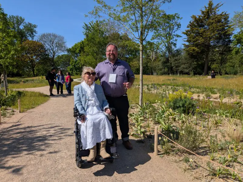Wheelchair user and companion stand on a wide garden path beside low planting and open meadow.