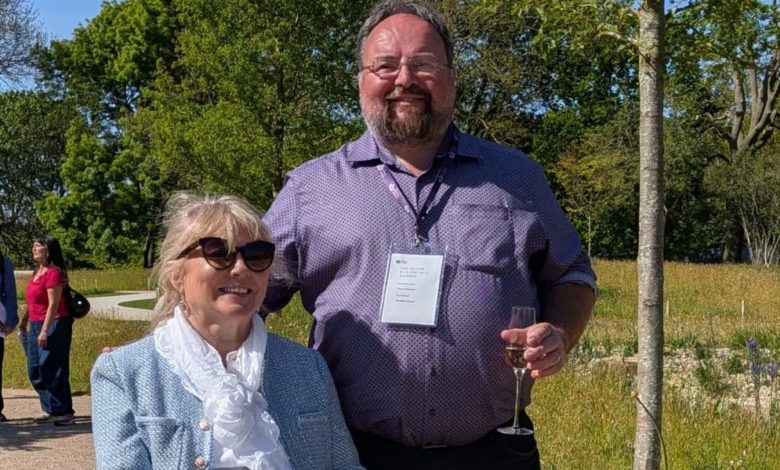 Wheelchair user and companion stand on a wide garden path beside low planting and open meadow.