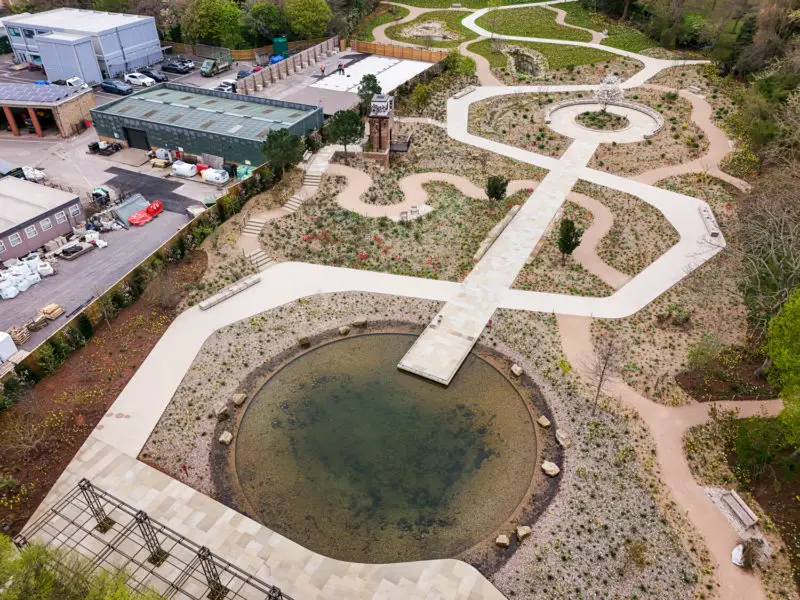Aerial view of a QE2 Garden garden with circular pond, wide paths, planted beds, and a water tower beside nearby buildings.