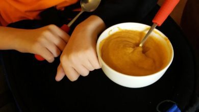 Child in an orange shirt sits with a bowl of orange soup on their lap, holding a spoon with a red handle.