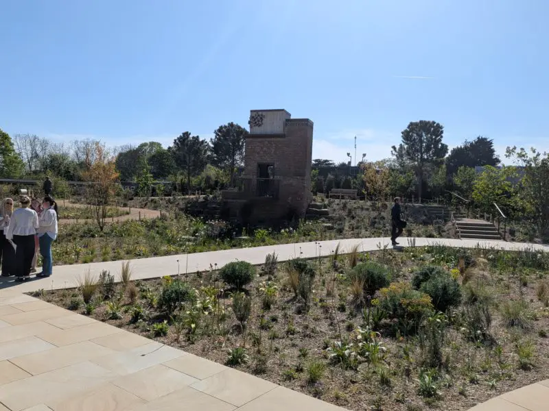 Curving path passes low planting grown in pale, stony soil, with the garden’s brick water tower ahead.