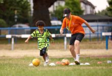 Child and football coach running drills with footballs and cones on a grass pitch during a community training session.