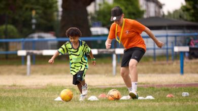 Child and football coach running drills with footballs and cones on a grass pitch during a community training session.