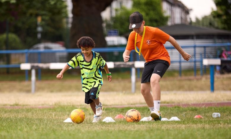 Child and football coach running drills with footballs and cones on a grass pitch during a community training session.