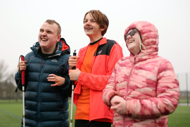 Three smiling young people wearing winter jackets stand outdoors holding white canes on a sports field.
