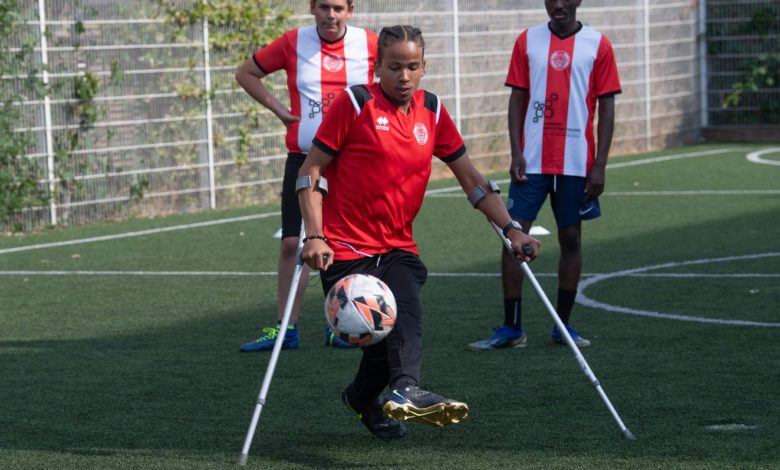 Teen on crutches kicks a soccer ball on an outdoor field while two teammates in red-and-white jerseys watch behind him.