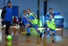 Children in blue soccer uniforms and green training bibs run after a bright green ball during an indoor soccer game while a coach watches from the side.