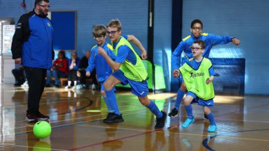 Children in blue soccer uniforms and green training bibs run after a bright green ball during an indoor soccer game while a coach watches from the side.