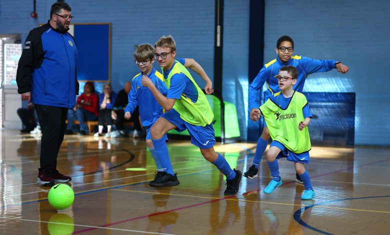 Children in blue soccer uniforms and green training bibs run after a bright green ball during an indoor soccer game while a coach watches from the side.