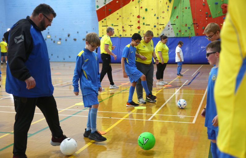 Children and coaches in blue and yellow sports uniforms practice soccer drills in an indoor gym with climbing walls in the background.
