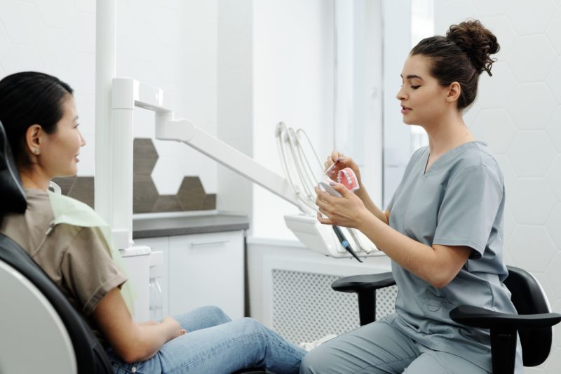 Dental professional explains orthodontic treatment using a model of teeth while speaking with a patient in a bright clinic.