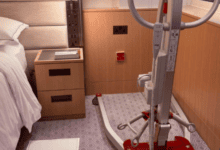 Mobile patient hoist positioned beside a hotel bed in an accessible guest room, with clear floor space for transfers.