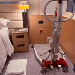 Mobile patient hoist positioned beside a hotel bed in an accessible guest room, with clear floor space for transfers.