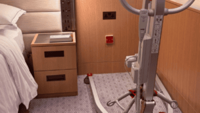 Mobile patient hoist positioned beside a hotel bed in an accessible guest room, with clear floor space for transfers.