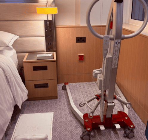 Mobile patient hoist positioned beside a hotel bed in an accessible guest room, with clear floor space for transfers.