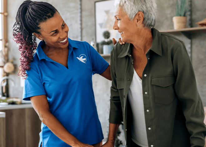 Caregiver in a blue uniform smiles while supporting an older woman standing in a bright, comfortable home.