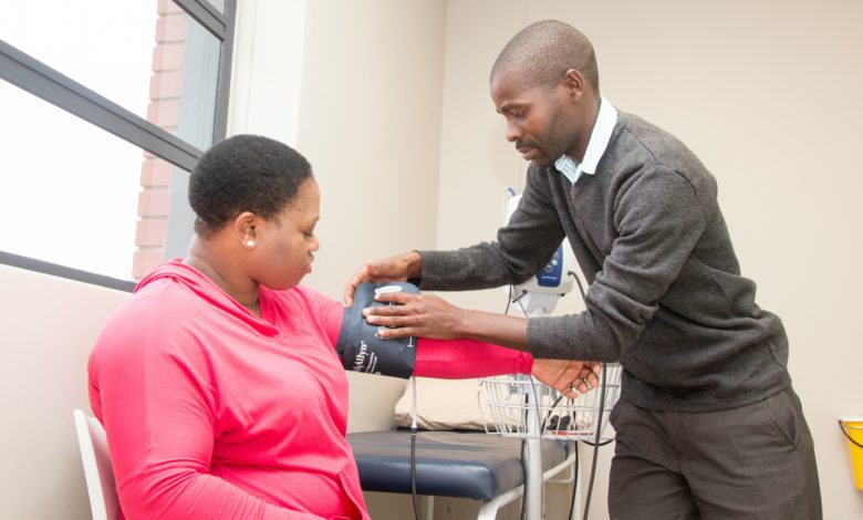 Healthcare worker measures a seated patient's blood pressure with a cuff in a bright examination room.