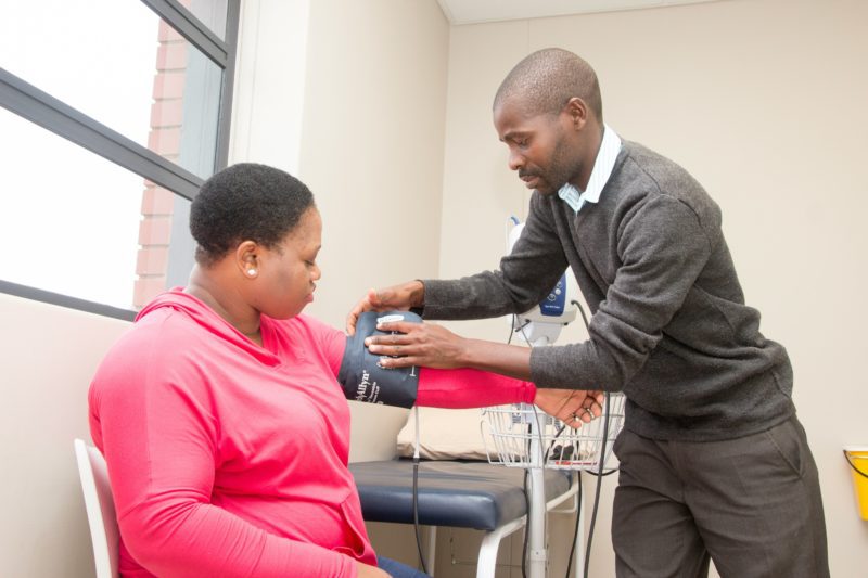 Healthcare worker measures a seated patient's blood pressure with a cuff in a bright examination room.