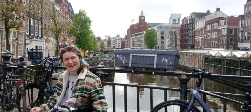 Liesl Hammer smiling by an Amsterdam canal, with bicycles, historic buildings and the waterway behind her.