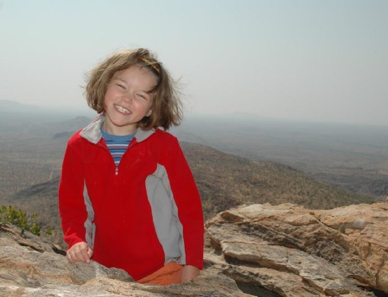 Smiling young Liesl Hammer wearing a red fleece, standing on rocky ground with a wide mountain landscape stretching into the distance.