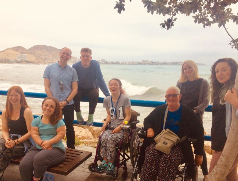 Liesl Hammer and a group of friends by the seaside, with Liesl and another wheelchair user smiling in the front row.