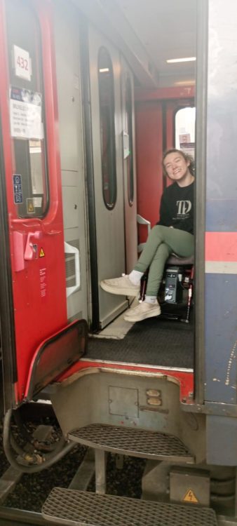 Liesl Hammer smiling from her wheelchair in the doorway of a train, train steps are shown, demonstrating that it wasn't step-free rail travel.