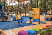 Children play in an inclusive sensory playground with a basket swing, accessible roundabout, musical play panel, rope climber, water play area and textured pathways while parents watch nearby.