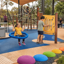 Children play in an inclusive sensory playground with a basket swing, accessible roundabout, musical play panel, rope climber, water play area and textured pathways while parents watch nearby.