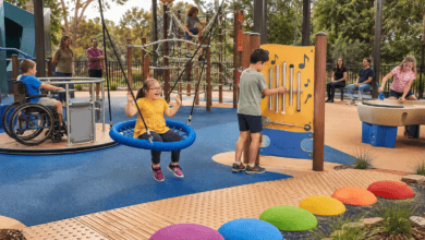 Children play in an inclusive sensory playground with a basket swing, accessible roundabout, musical play panel, rope climber, water play area and textured pathways while parents watch nearby.