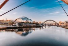 Panoramic view of the River Tyne with the Tyne Bridge and The Glasshouse reflected in calm water beneath a blue sky.