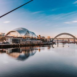 Panoramic view of the River Tyne with the Tyne Bridge and The Glasshouse reflected in calm water beneath a blue sky.