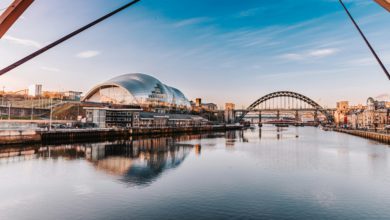 Panoramic view of the River Tyne with the Tyne Bridge and The Glasshouse reflected in calm water beneath a blue sky.
