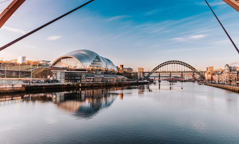 Panoramic view of the River Tyne with the Tyne Bridge and The Glasshouse reflected in calm water beneath a blue sky.