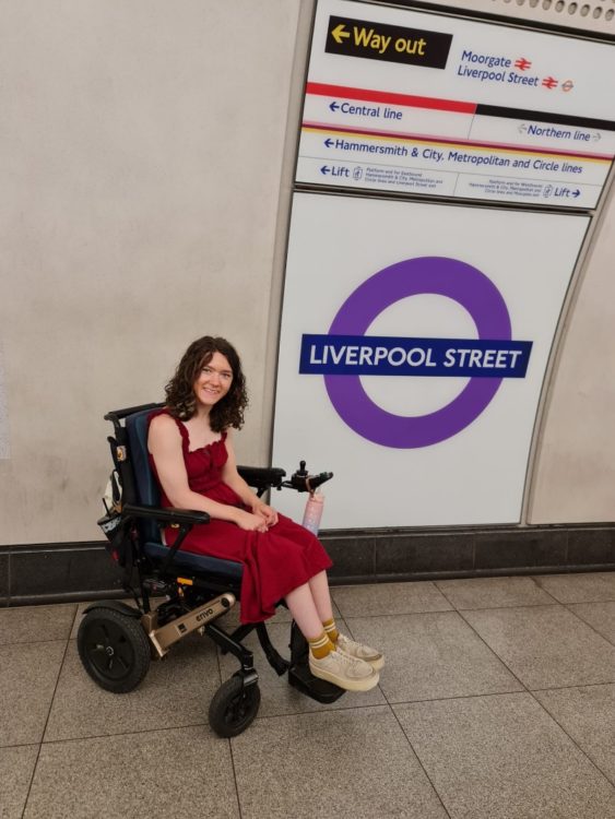 Liesl Hammer in her electric wheelchair beside a Liverpool Street station sign in London.