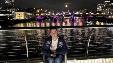 Liam Virgo in his wheelchair on a footbridge over the Thames at night, with Tower Bridge, the Shard and a full moon behind him.