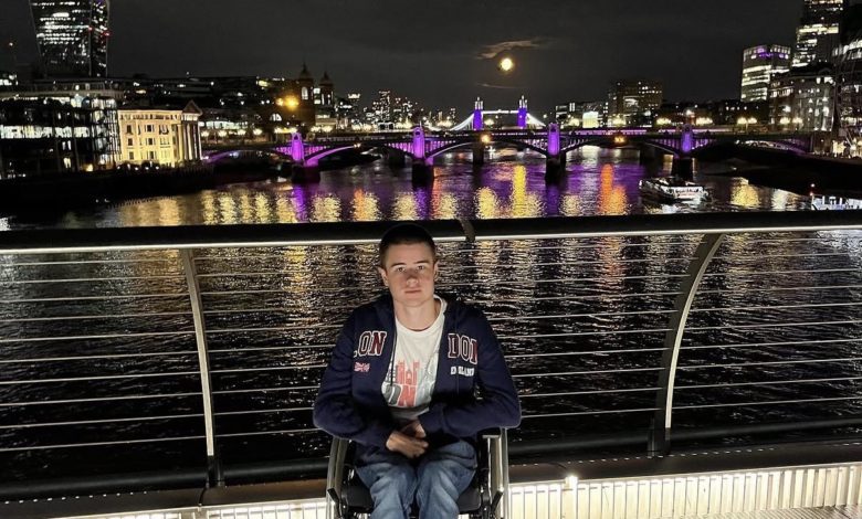 Liam Virgo in his wheelchair on a footbridge over the Thames at night, with Tower Bridge, the Shard and a full moon behind him.