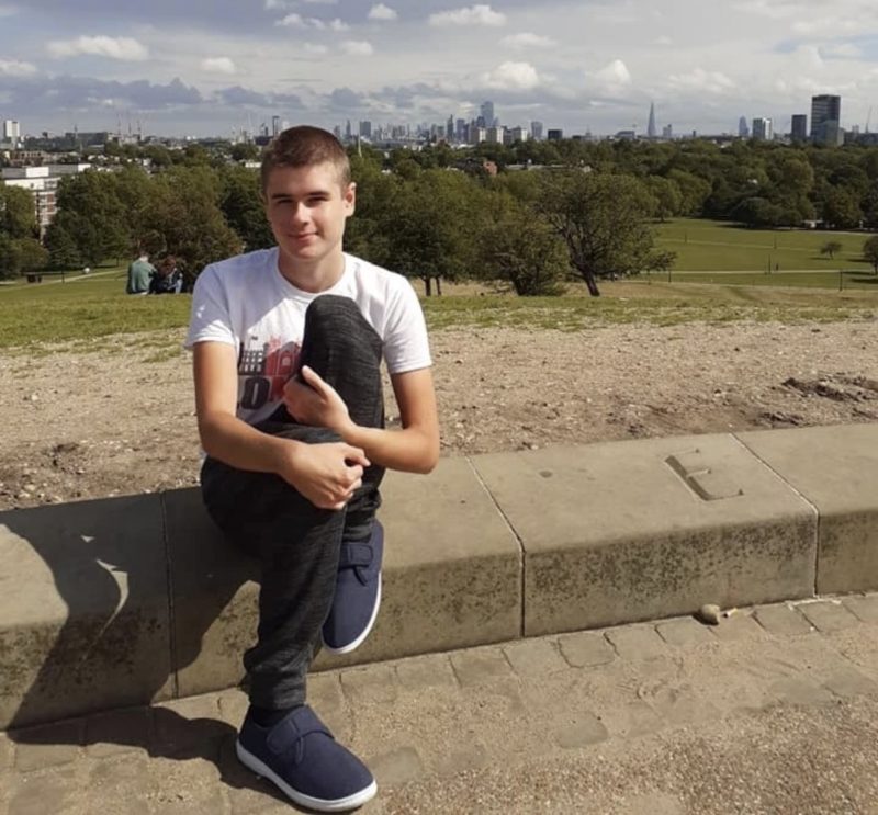 Liam as a teenager sitting outdoors in a London park, with the city skyline visible in the distance.
